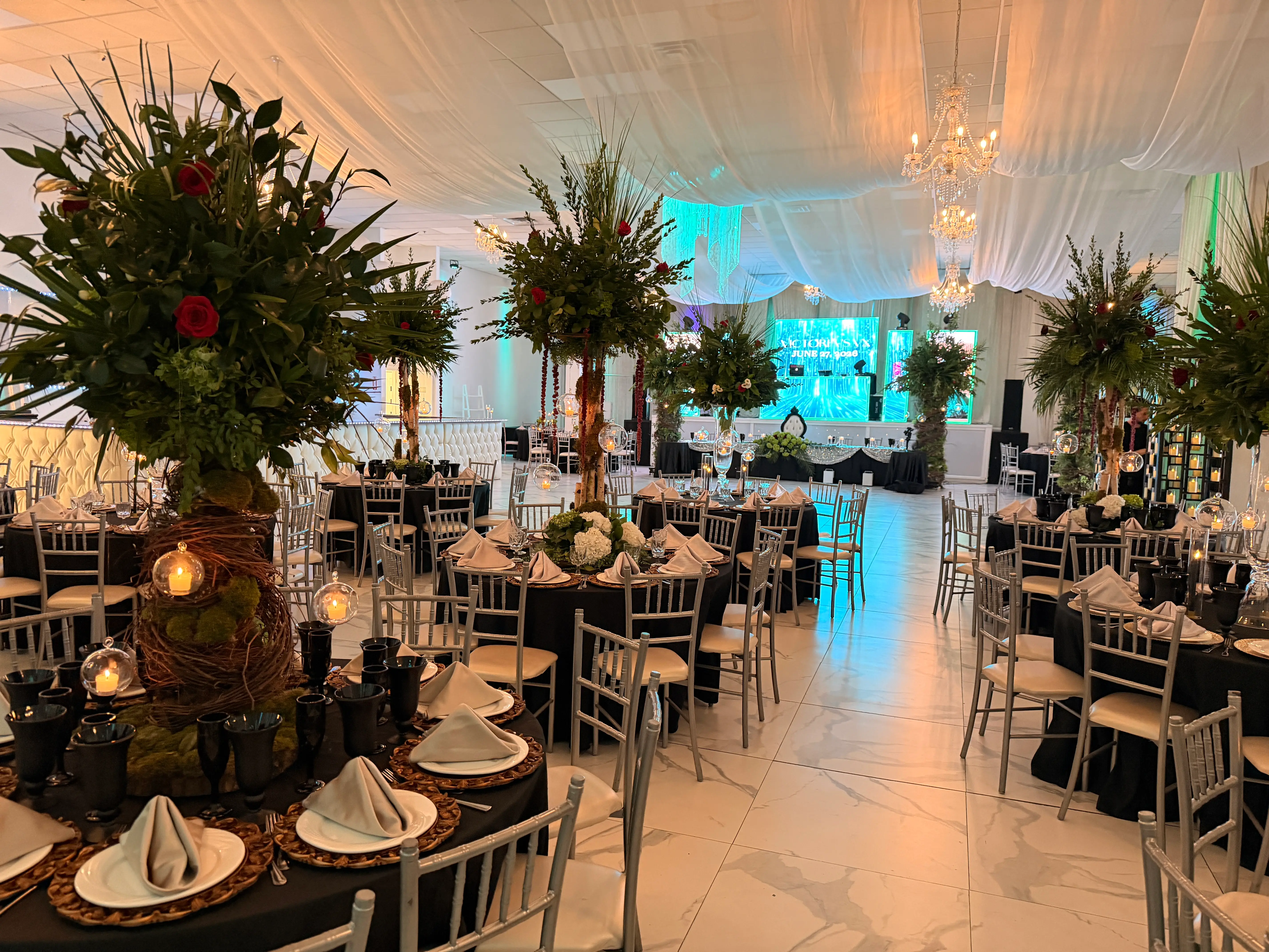 Wide view of the ballroom — white draped ceiling, chandelier, marble dance floor, and tables dressed in black with greenery centerpieces