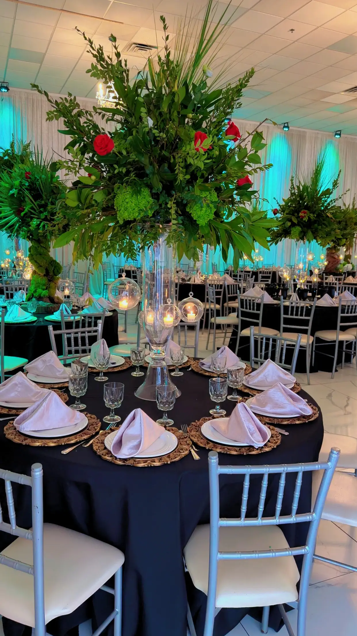 Elegant table in black linen with gold chargers, white napkins, and a tall greenery centerpiece with hanging glass candle orbs
