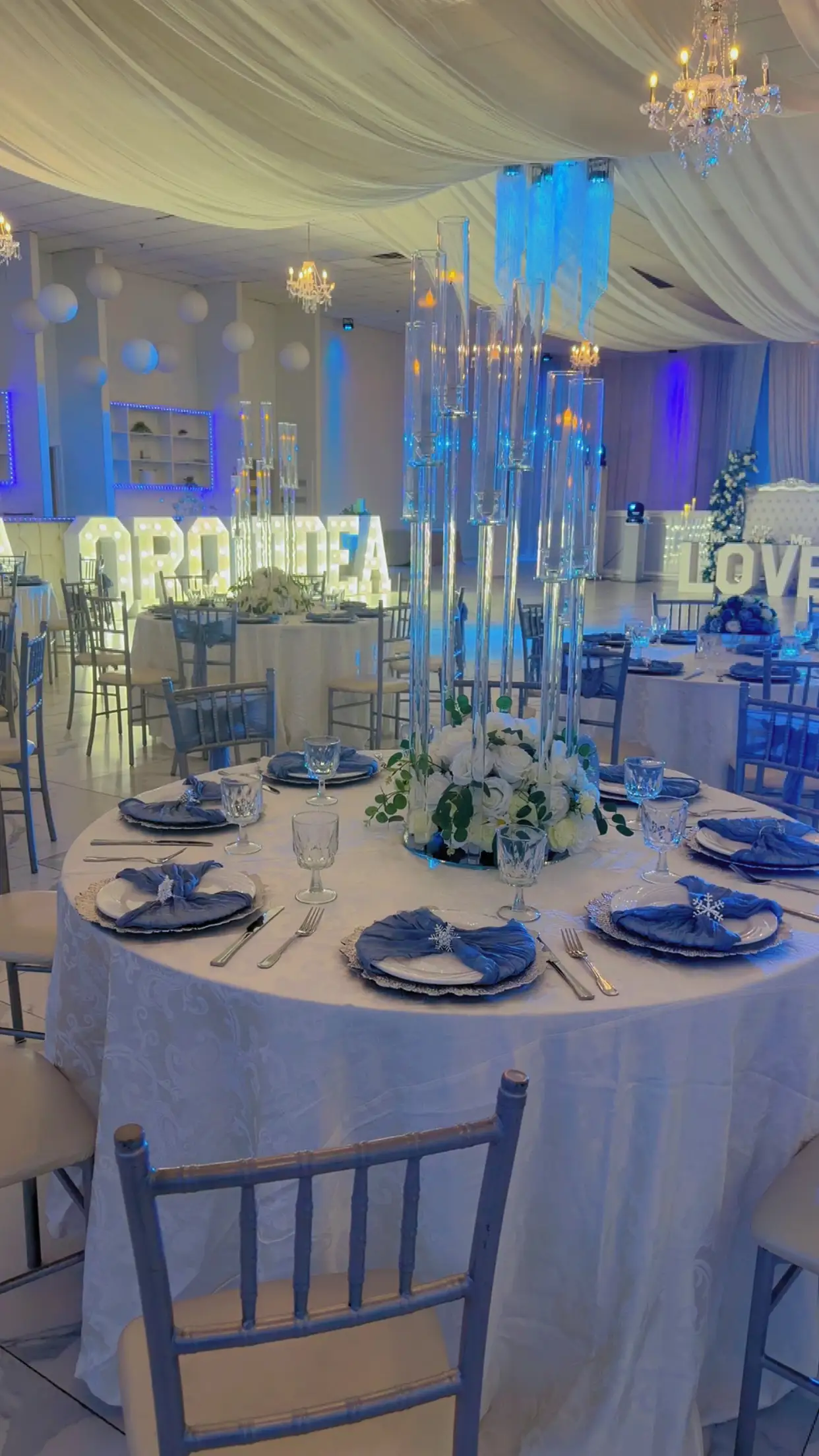 Round reception table with blue napkins, white floral centerpiece, and tall glass candlesticks, marquee letters glowing in the background