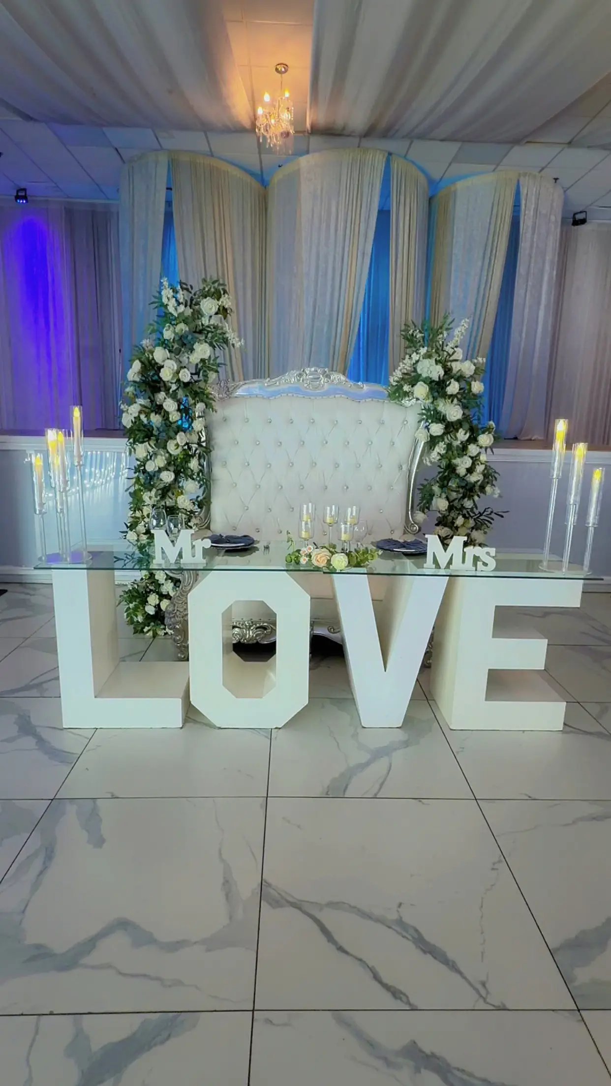 Wedding sweetheart table with a white tufted loveseat, Mr & Mrs sign, marquee LOVE letters, floral arch, and candlesticks